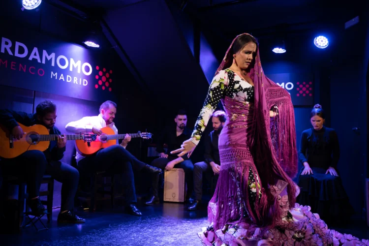 Primer plano de una bailaora de flamenco en plena actuación en el escenario de Cardamomo. La iluminación es de un rojo intenso y dramático sobre un fondo oscuro. Ella viste un traje tradicional oscuro y se encuentra en una postura dinámica y llena de fuerza, con una expresión de profunda pasión y entrega.