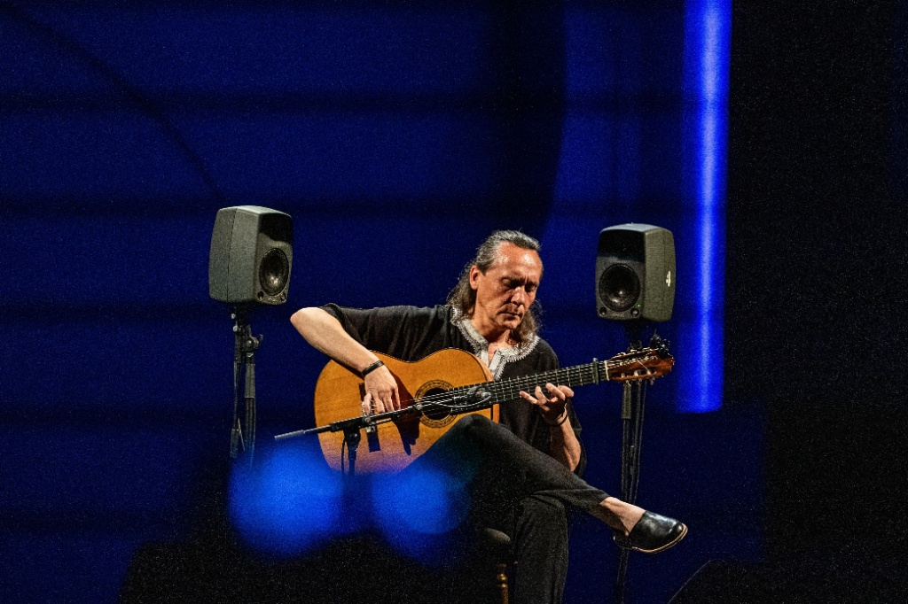 Fotografía de Vicente Amigo tocando la guitarra flamenca en un escenario con iluminación azul. Está sentado con las piernas cruzadas, vestido de oscuro y tocando con profunda concentración junto a dos monitores de sonido.