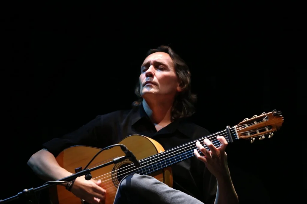 Fotografía del guitarrista flamenco Vicente Amigo tocando la guitarra en directo sobre el escenario. Aparece con los ojos cerrados y gesto de profunda concentración, vistiendo una camisa negra sobre un fondo oscuro.