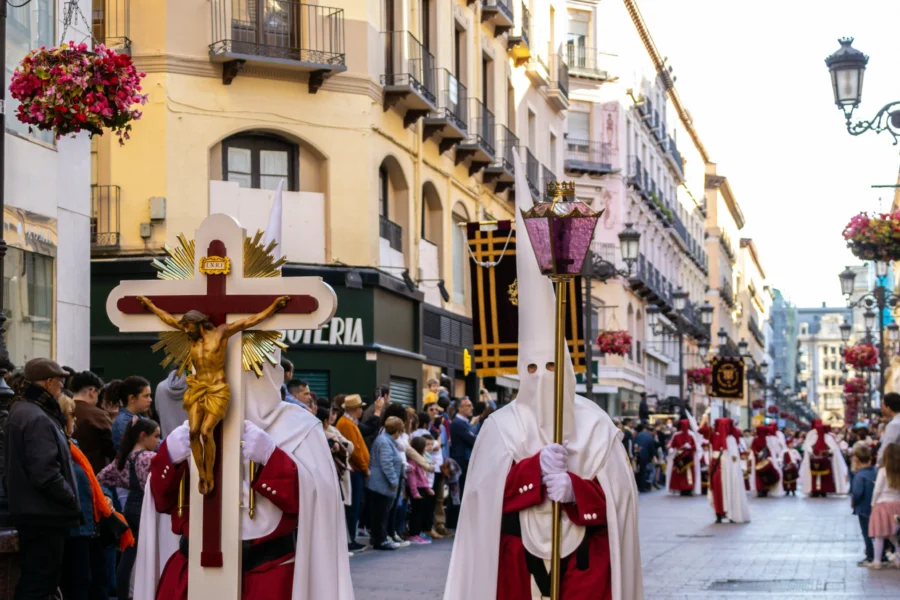 Fotografía de una procesión de Semana Santa en una calle histórica de Madrid con edificios clásicos. En primer plano, dos nazarenos con capirotes blancos y capas rojas. El de la izquierda porta un gran crucifijo de madera. El de la derecha lleva un cetro metálico coronado con un relicario de cristal rosa. Una multitud de espectadores observa desde las aceras.