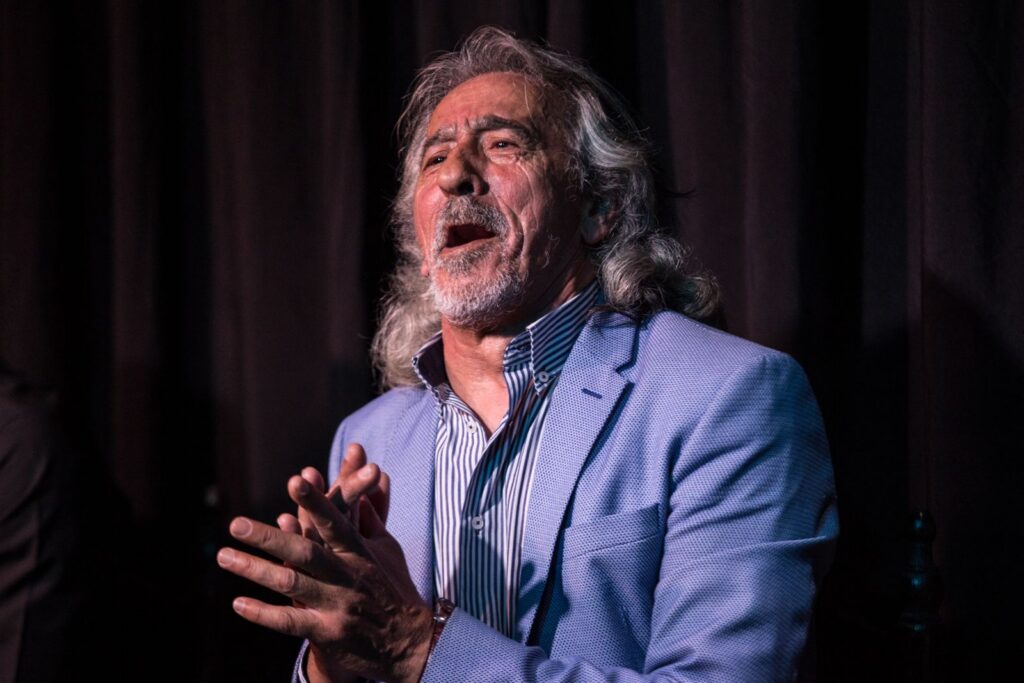 Fotografía del cantaor flamenco Capullo de Jerez actuando. Es un hombre mayor con pelo largo y canoso y barba, captado en un momento de canto apasionado con la boca abierta y la mirada hacia arriba. Viste una chaqueta azul claro texturizada sobre una camisa de rayas azules y blancas, y mantiene las manos entrelazadas a la altura del pecho frente a un fondo de escenario oscuro.