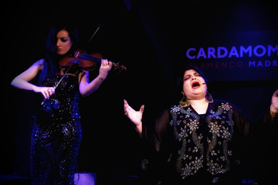 Fotografía en directo de una cantaora de flamenco interpretando con gran emoción y los ojos cerrados. A su lado, un músico la acompaña tocando el violín, ilustrando la fusión flamenca. En el fondo oscuro del escenario destaca el logotipo iluminado en letras rojas del tablao Cardamomo.