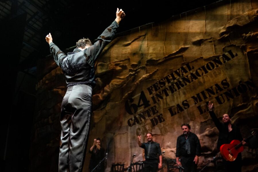 Ballerino di flamenco di spalle sul palco durante il Festival Internazionale del Cante de las Minas a La Unión, con musicisti che incitano sullo sfondo.
